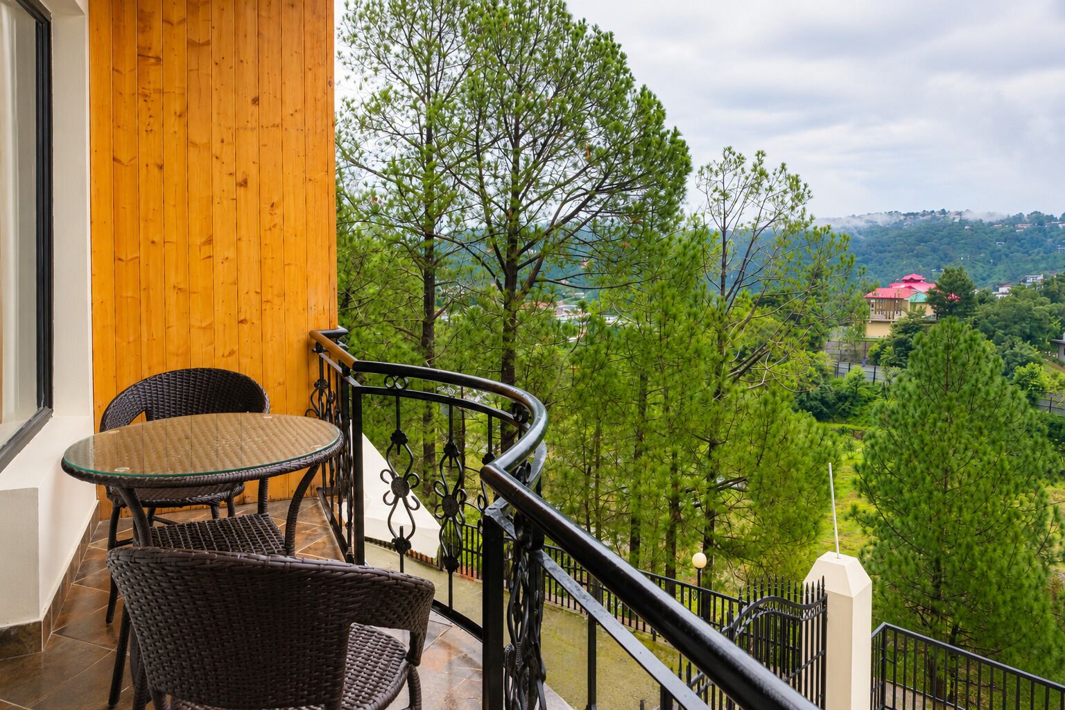 A balcony with wicker furniture, a view of a green landscape, and a distant building with a red roof.
