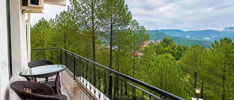 A balcony with a glass table and wicker chairs, overlooking a forested area with distant buildings.