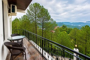 A balcony with a glass table and wicker chairs, overlooking a forested area with distant buildings. - Hotel Heaven & Hills Kasauli (Kasauli)
