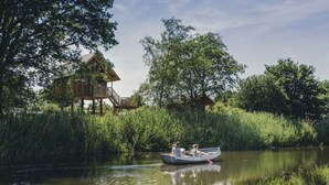 A wooden house with a deck, surrounded by lush greenery and a calm river. - Charming Safari Tent for Family Glamping (Zuna)