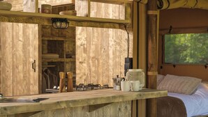 A rustic wooden cabin interior with a kitchenette, a bed, and a window overlooking greenery. - Charming Safari Tent for Family Glamping (Zuna)