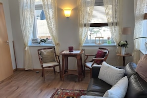 Ein gemütliches Wohnzimmer mit Ledersofa, Holztisch mit Stühlen, einem kleinen Beistelltisch mit Lampe und einem Fenster mit Vorhängen.
