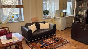 Ein Wohnzimmer mit einem dunkelbraunen Ledersofa, einem gemusterten Teppich, einem Esstisch mit einem violetten Kerzenlicht und einer Küchenzeile mit Kühlschrank und Schränken.