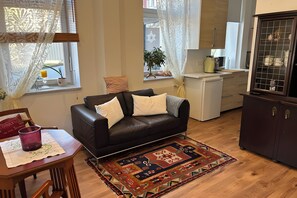 Una sala de estar con un sofá de cuero oscuro, un tapete estampado, una mesa de comedor con una vela morada y una zona de cocina con un refrigerador y armarios.