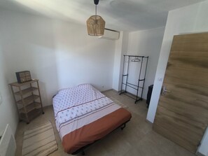 A bedroom with a bed, a wooden wardrobe, a hanging clothes rack, a small table with a lamp, and a ceiling fan.