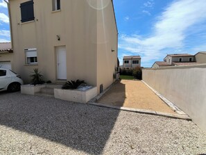 Un immeuble résidentiel avec une allée en gravier, une voiture blanche garée sur le côté gauche et un petit jardin avec des plantes.
