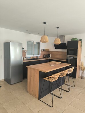 Une cuisine moderne avec un réfrigérateur en acier inoxydable, un îlot central en bois, des meubles de cuisine noirs et des chaises en osier.