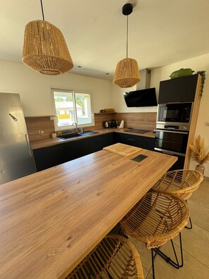 A modern kitchen with a wooden island, hanging wicker pendant lights, a built-in oven, and a large window. - 3-bedroom villa with garage, covered terrace and air-conditioned pétanque court (Raphele les Arles)