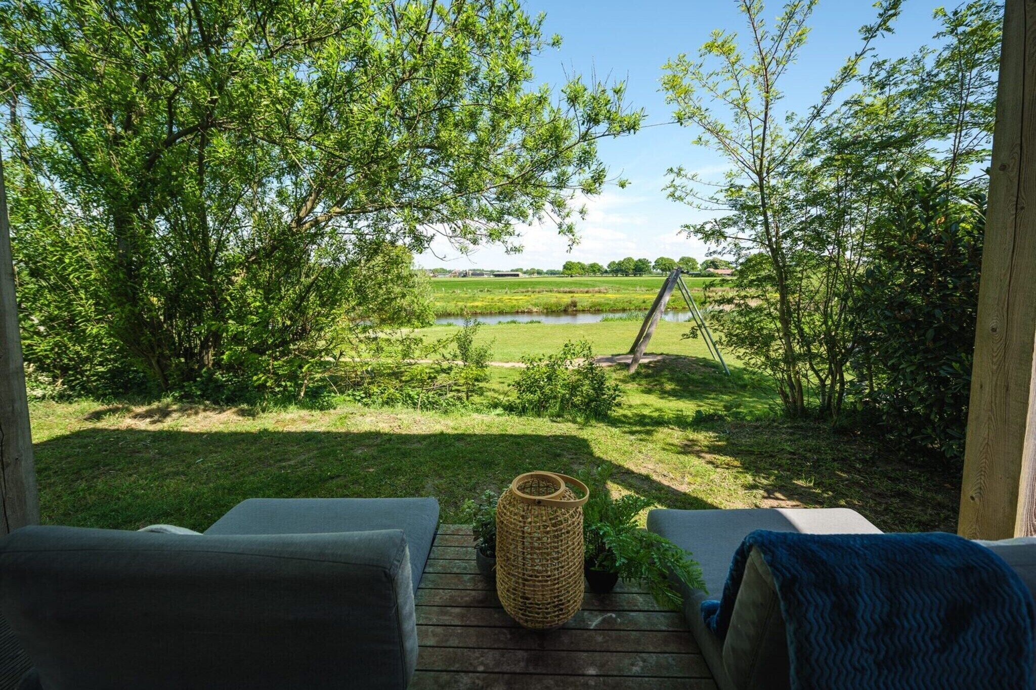 A deck with two sofas, a wicker basket, and a view of a grassy field with trees and a body of water.