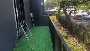 Ein Balkon mit Kunstgrashalm, einer Metallleiter und Blick auf geparkte Autos und Grünflächen.