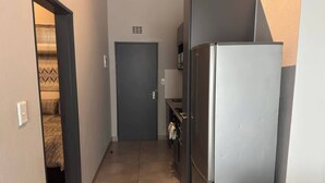 Un pasillo estrecho con una puerta al final, un refrigerador y una pequeña cocina con un lavabo y un perchero.