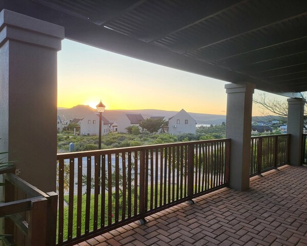A covered balcony with a brick floor, a metal railing, and a view of houses and greenery in the distance.