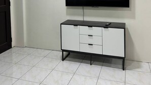 Een kamer met een flatscreen televisie aan de muur, een witte kast met lades en een betegelde vloer.