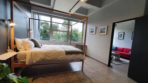 Ein Schlafzimmer mit einem Himmelbett, einer Nachttischlampe, einem Fenster mit Blick auf Bäume und ein rotes Sofa im angrenzenden Raum.
