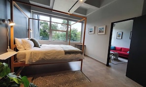 Ein Schlafzimmer mit einem Himmelbett, einer Nachttischlampe, einem Fenster mit Blick auf Bäume und ein rotes Sofa im angrenzenden Raum.