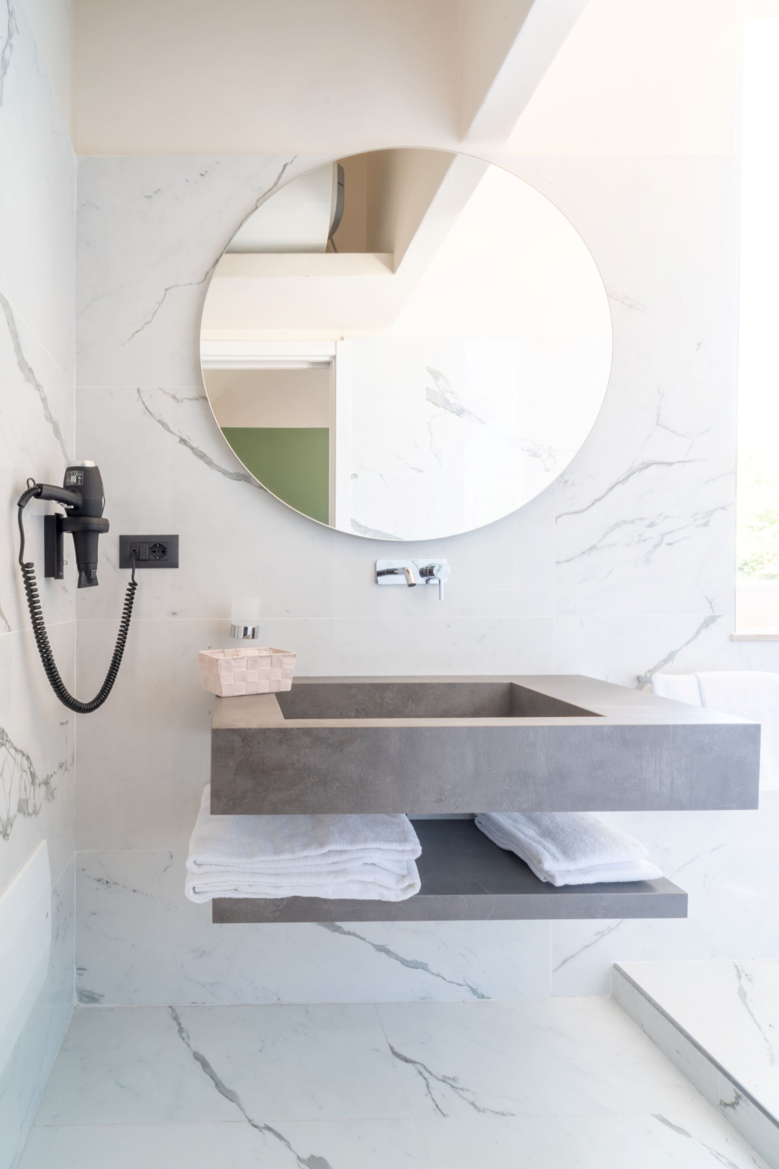 A bathroom with a marble wall, a round mirror, a hairdryer, a towel rack, and a sink with a shelf.