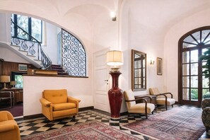 Un vestíbulo amplio con piso a cuadros, una escalera con barandilla ornamentada y un gran ventanal con celosía metálica decorativa.