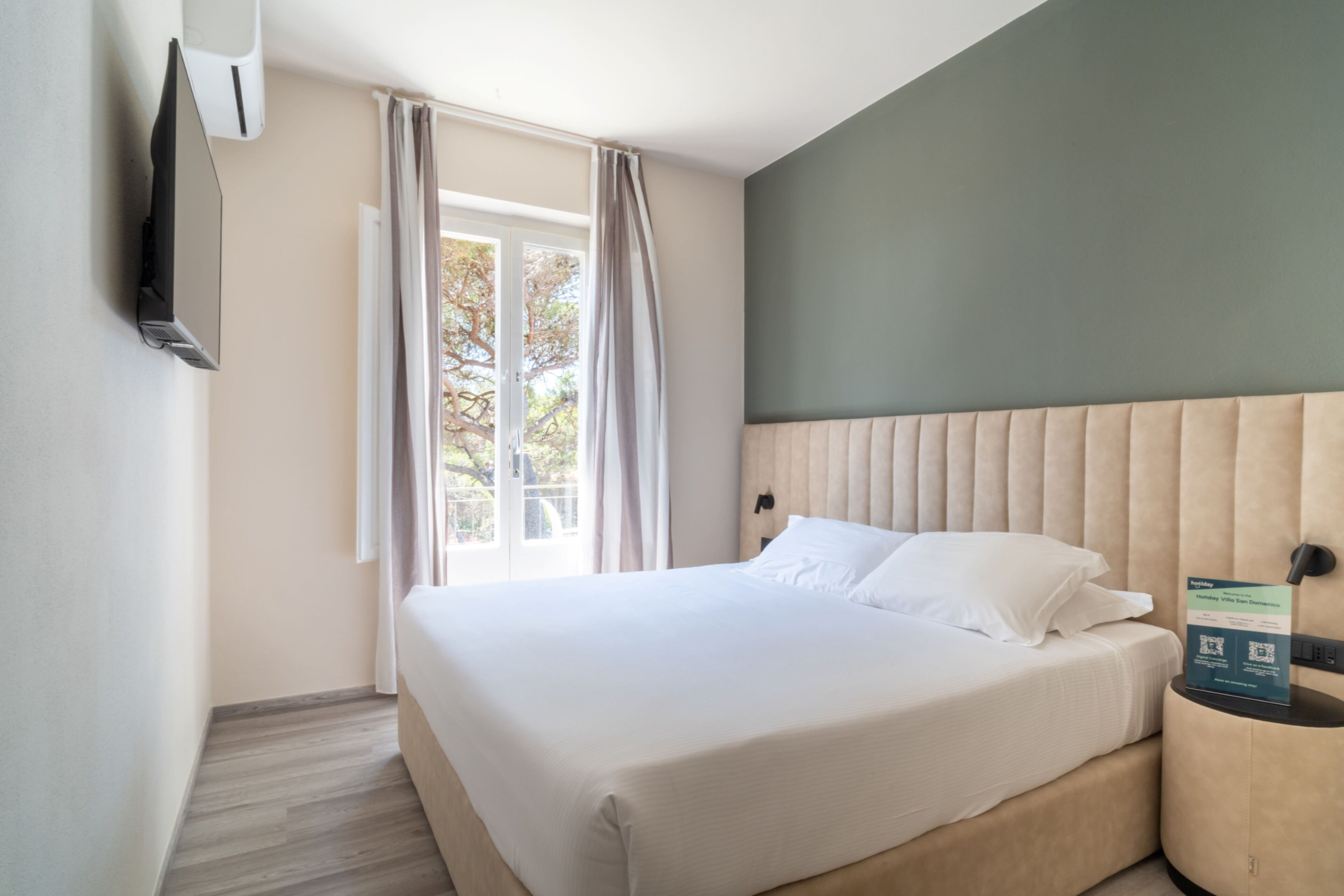 A hotel room with a bed, a TV mounted on the wall, a bedside table, and a window with curtains.