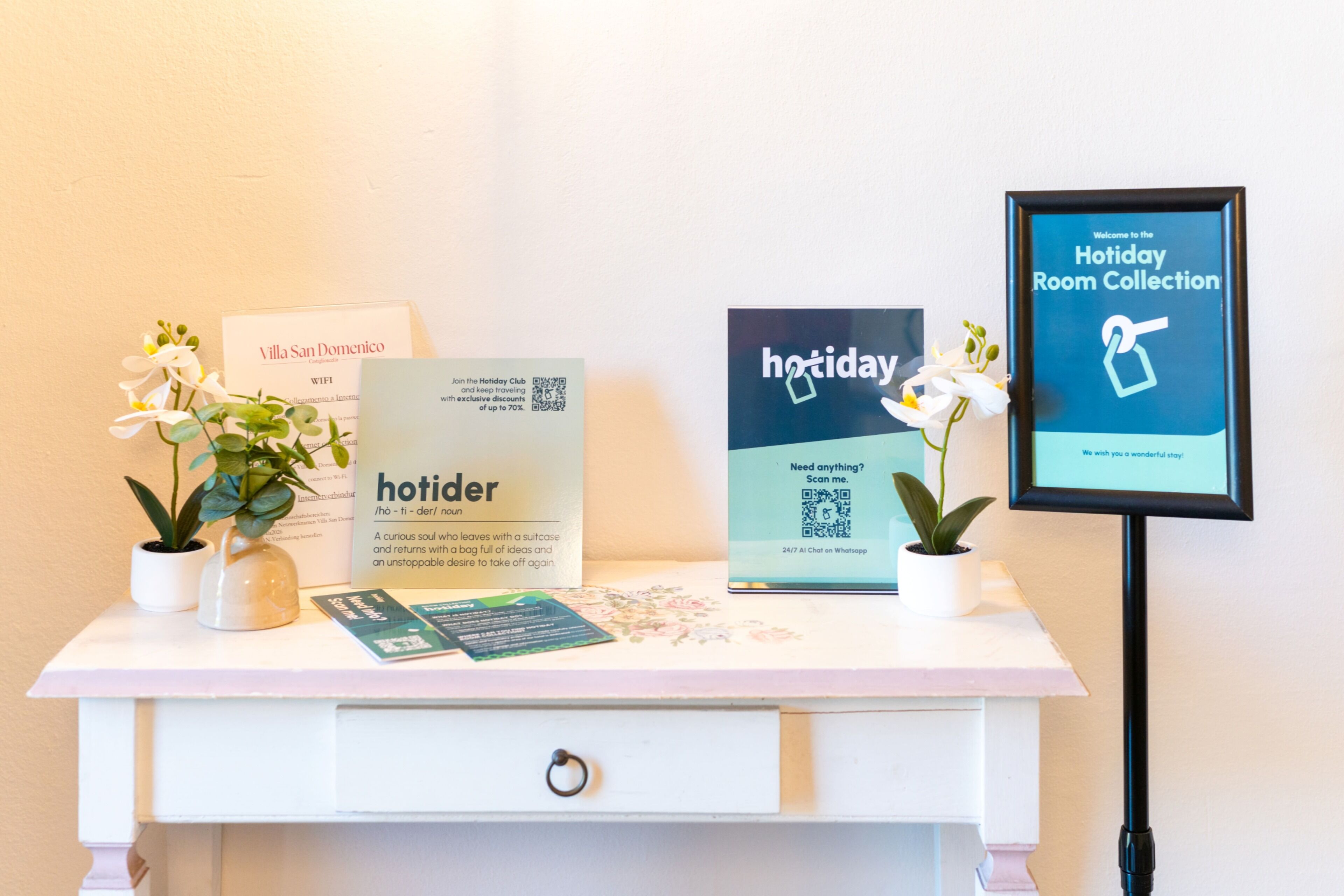 A reception desk with a sign for 'Villa San Domenico' and a 'hotider' sign, a QR code, and a framed welcome sign for the 'Holiday Room Collection'.