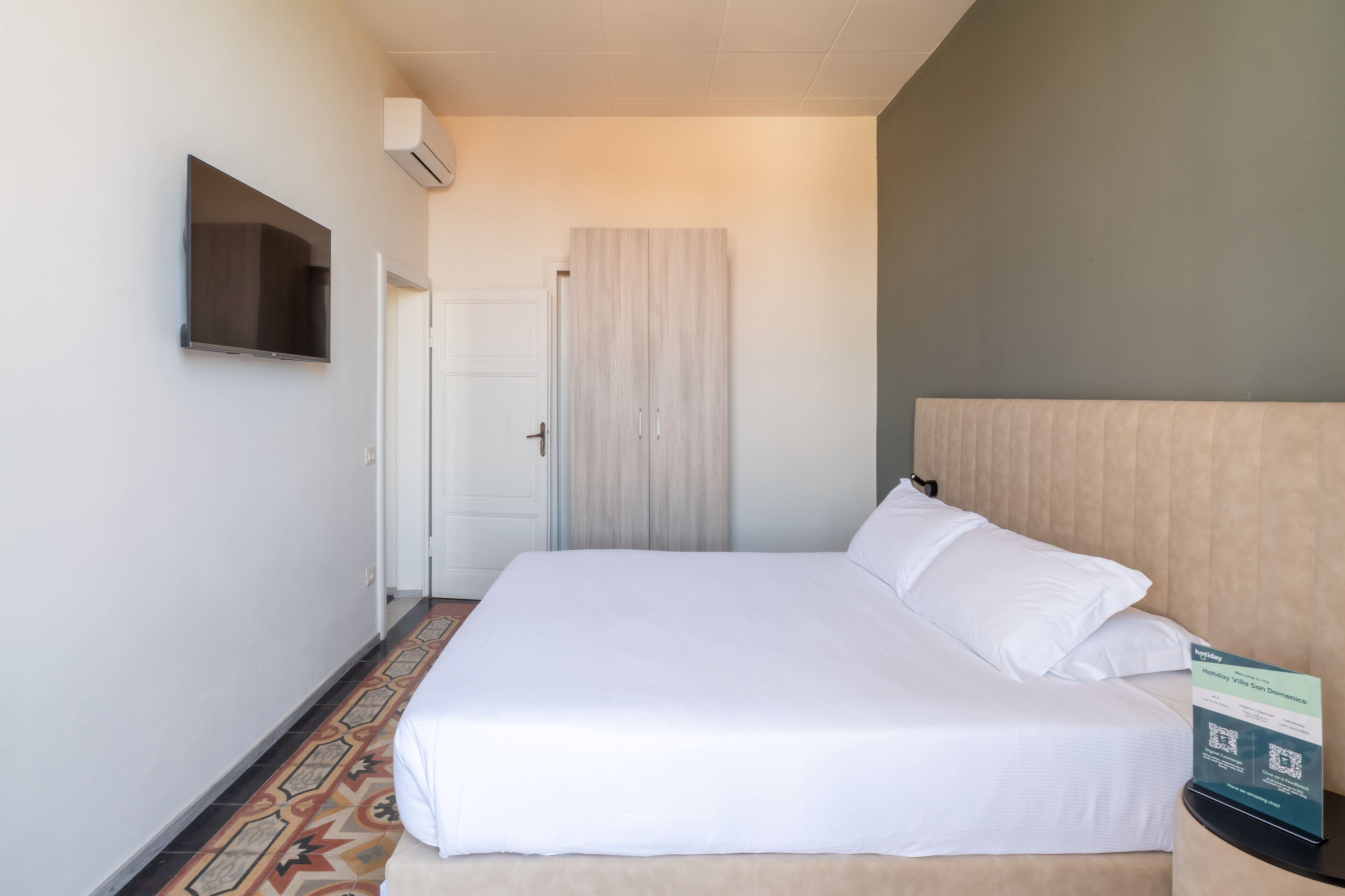 A hotel room with a bed, a flat-screen TV mounted on the wall, a door, and a sign on the nightstand.