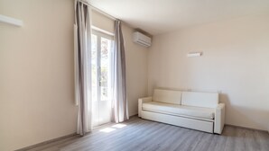 A room with a light-colored wooden floor, a white sofa, a sliding glass door with curtains, and an air conditioning unit mounted on the wall. - Hotiday Villa San Domenico (Rosignano Marittimo)