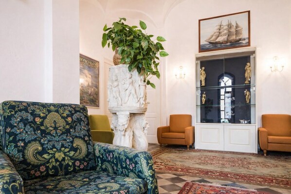 Una habitación con un sofá estampado, dos sillones, un pilar de mármol con una planta, una vitrina de cristal con esculturas y un cuadro enmarcado de un barco.