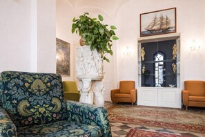 Una habitación con un sofá estampado, dos sillones, un pilar de mármol con una planta, una vitrina de cristal con esculturas y un cuadro enmarcado de un barco.