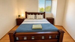 A neatly made bed with a blue blanket and pillows, wooden headboard, bedside tables with lamps, and a window with blinds.