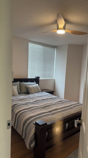 Un dormitorio con cama, mesita de noche, ventana con persianas y ventilador de techo.