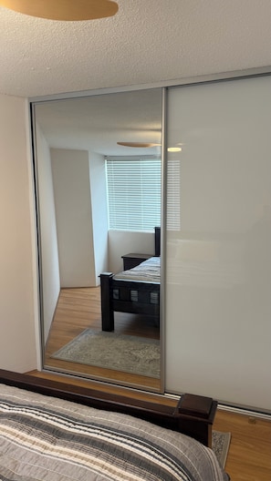 A bedroom with a bed, a bedside table, a window with blinds, and a mirror. - Modern 2BR Apartment with Free Parking | Near Perth CBD & Beaches. (Wembley)