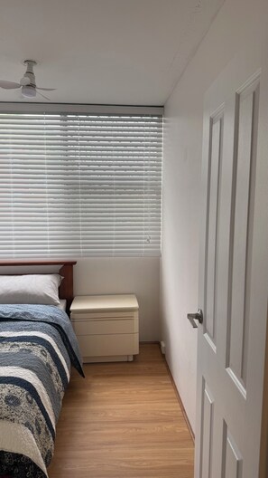 Un dormitorio con una cama, una mesita de noche, una ventana con persianas, un ventilador de techo y una puerta.