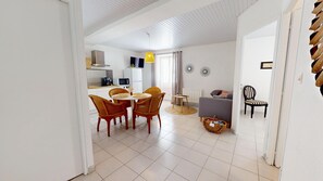 A modern kitchen with white cabinets, a dining area with wicker chairs, a grey sofa, and a black chair with striped cushions. - HAUBAN - Charm & Authenticity in the Old Port (Saint-Gilles-Croix-de-Vie)