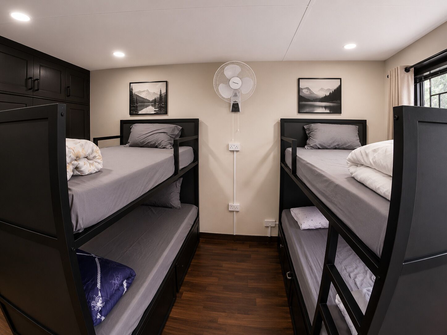 A dormitory room with bunk beds, a wall-mounted fan, and two landscape paintings.