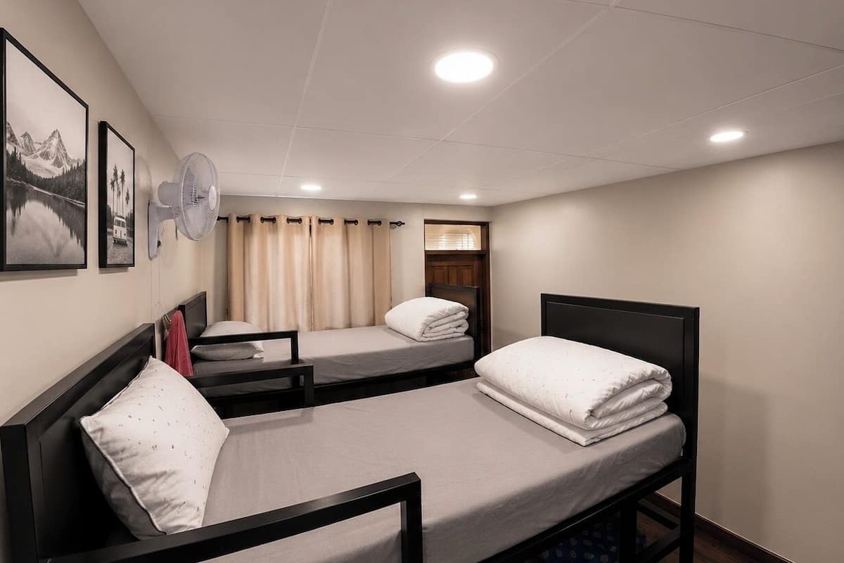 A dormitory room with two beds, a wall-mounted fan, and a window with curtains.