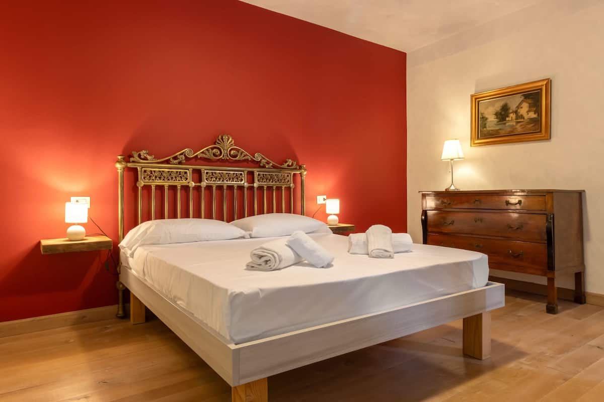 Uma cama com cabeceira de madeira, duas luminárias de cabeceira, uma cômoda de madeira e um quadro na parede.