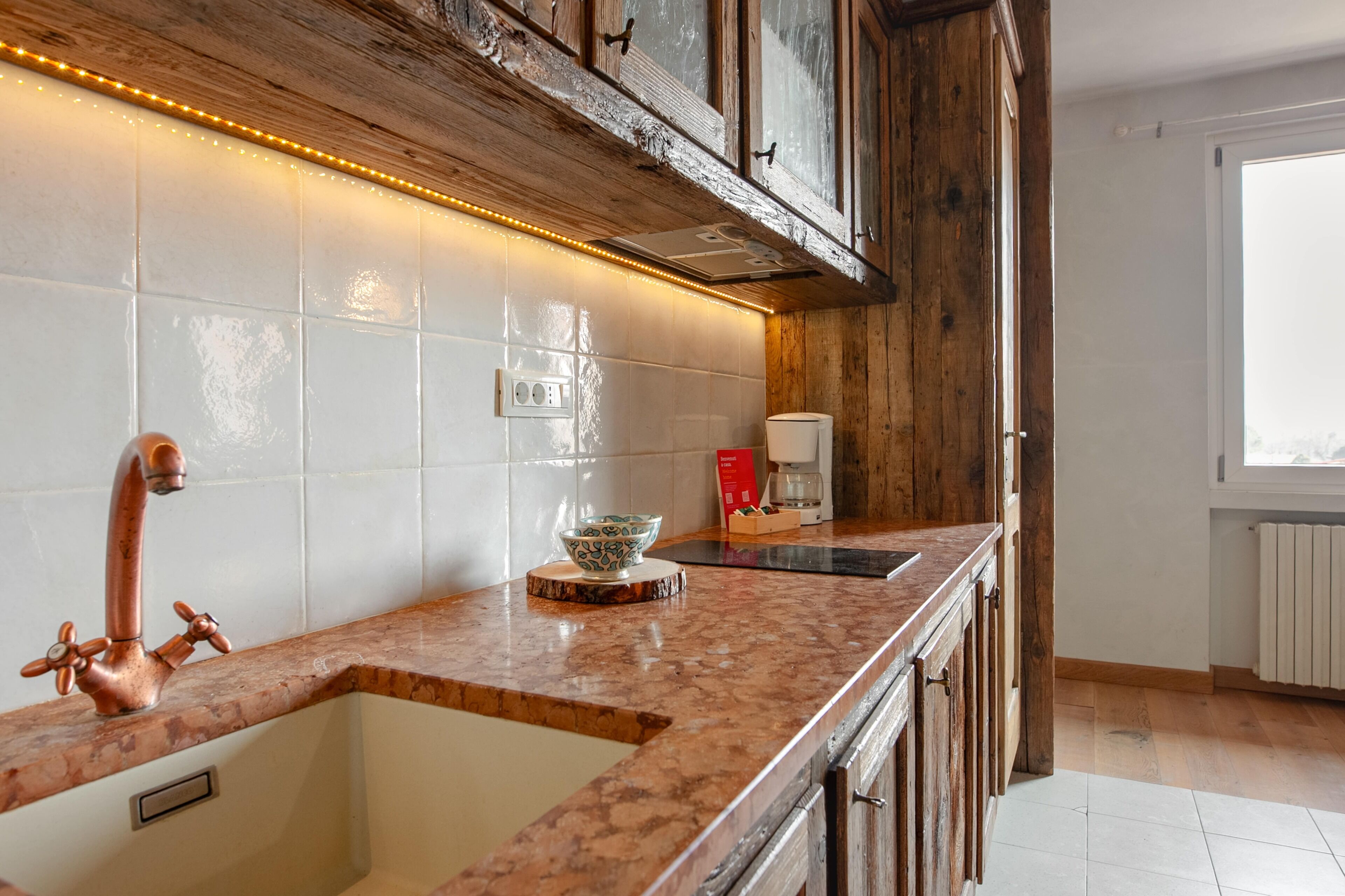Een keuken met een koperen kraan, een witte betegelde spatwand, houten kastjes en een raam met uitzicht.