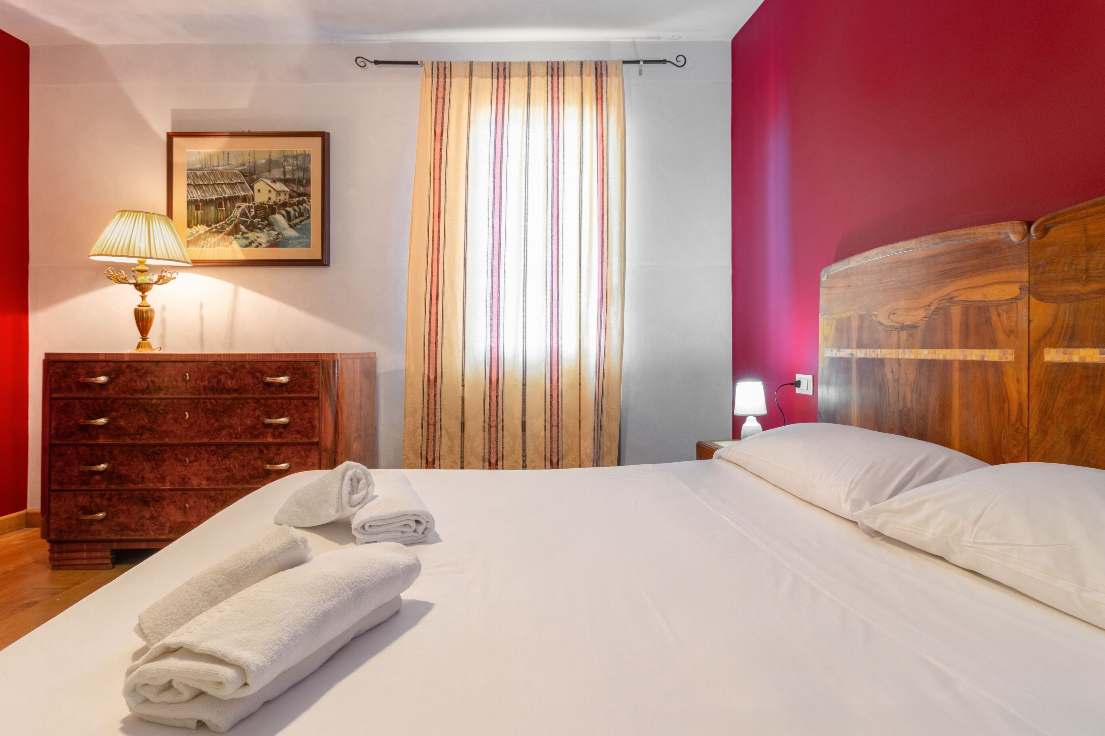 Cama bem arrumada com lençóis brancos, cômoda de madeira com gavetas, luminária, quadro pendurado na parede e cortina listrada.