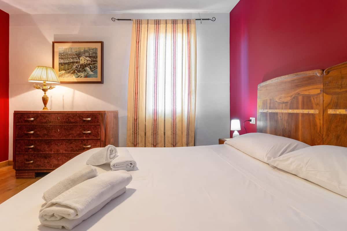 Cama bem arrumada com lençóis brancos, cômoda de madeira com gavetas, luminária, quadro pendurado na parede e cortina listrada.