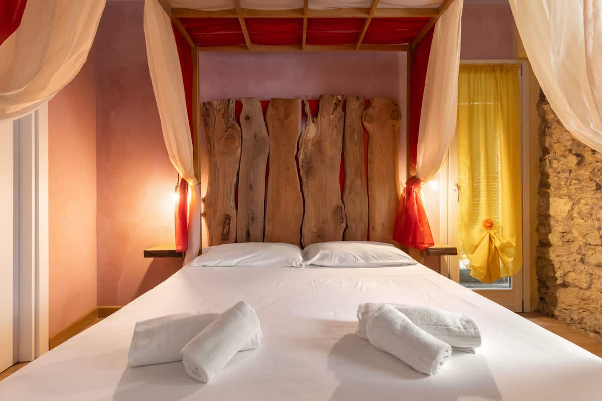 Cama com lençóis brancos, duas toalhas, cabeceira de madeira e cortinas vermelhas e amarelas.