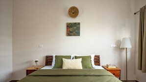 A neatly made bed with a green bedspread, two pillows, a small bedside table with a vase of flowers, a wall-mounted clock, and a lamp.