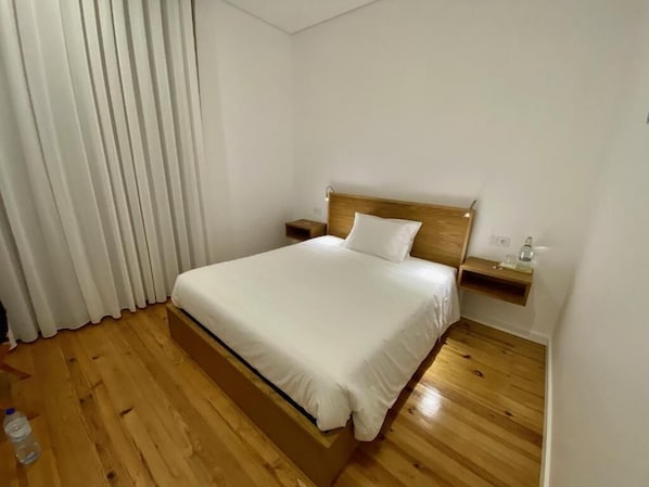 A bedroom with a wooden bed, white bedding, a nightstand, and a window with white curtains.
