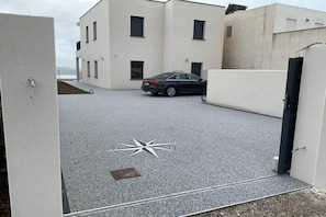 Ein modernes Gebäude mit einem geparkten Auto davor, einem Kompass-ähnlichen Design im Boden und Blick auf das Meer.