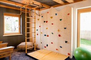 Eine Kletterwand mit einer Holzleiter, einer gepolsterten Bank und einem grünen Trainingsball in einem Raum mit Fenster und Tür.