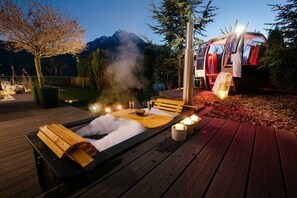 Ein Whirlpool auf einer Holzterrasse mit einer Bank und Kerzen, umgeben von Bäumen und einem Berg im Hintergrund.