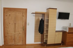 Ein Zimmer mit Holztür, einem Kleiderschrank mit offenen Regalen, einem an der Wand befestigten Fernseher und einem Tisch mit Stoffüberzug.