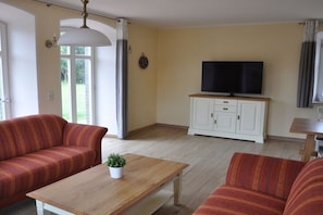 Ein Wohnzimmer mit einer Couch, einem Couchtisch, einem Fernseher auf einem Schrank und einem Fenster mit Vorhängen.