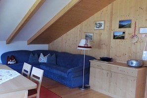 Ein Zimmer mit Holzboden, einem blauen Sofa, einem Tisch mit weißem Tischlein und einem Schrank mit Radio.
