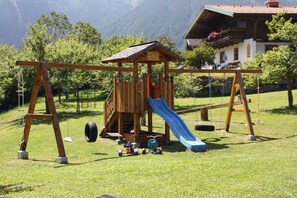 A wooden play structure with a slide, swings, and a tire swing in a grassy area with trees and a building in the background. - Holiday home in Krimml with swimming pool (Krimml)