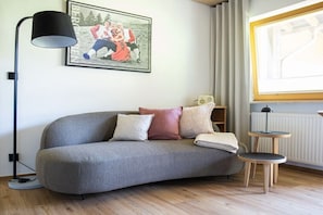 A modern living room with a curved grey sofa, a floor lamp, a small round table, a framed family photo, and a window with curtains. - Apartment in Mittelberg with swimming pool (Mittelberg)