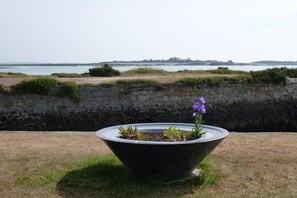 Ein großer Pflanzkasten mit Pflanzen und einem violetten Strauch im Vordergrund, eine Steinmauer und ein Blick auf ein Gewässer und entfernte Landschaft.